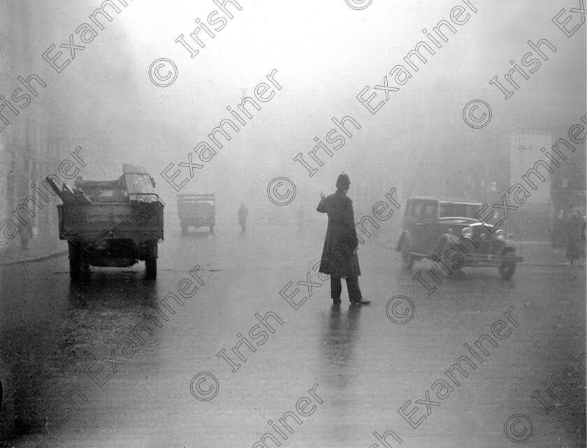 823344 
 For 'READY FOR TARK'
A Garda attempts to direct traffic in heavy fog at Singer's Corner, Cork 04/01/1934 Ref. 240B Old black and white cars winter weather smog police